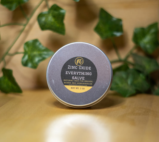 Zinc Oxide Everything Salve tin on a wooden surface with green leaves in the background